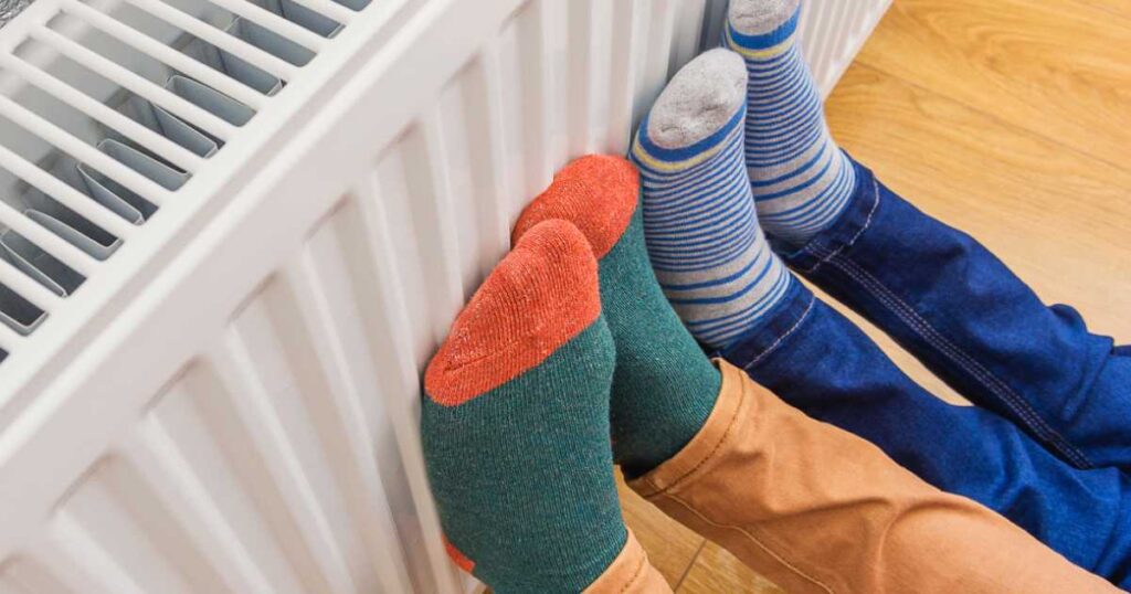 Adult and child wearing colorful pair of woolly socks warming cold feet in front of heating radiator. Gas heater at home.