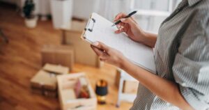 One woman standing in her home with boxes and packing supplies in the background and checking the list of her stuff and moving tasks.